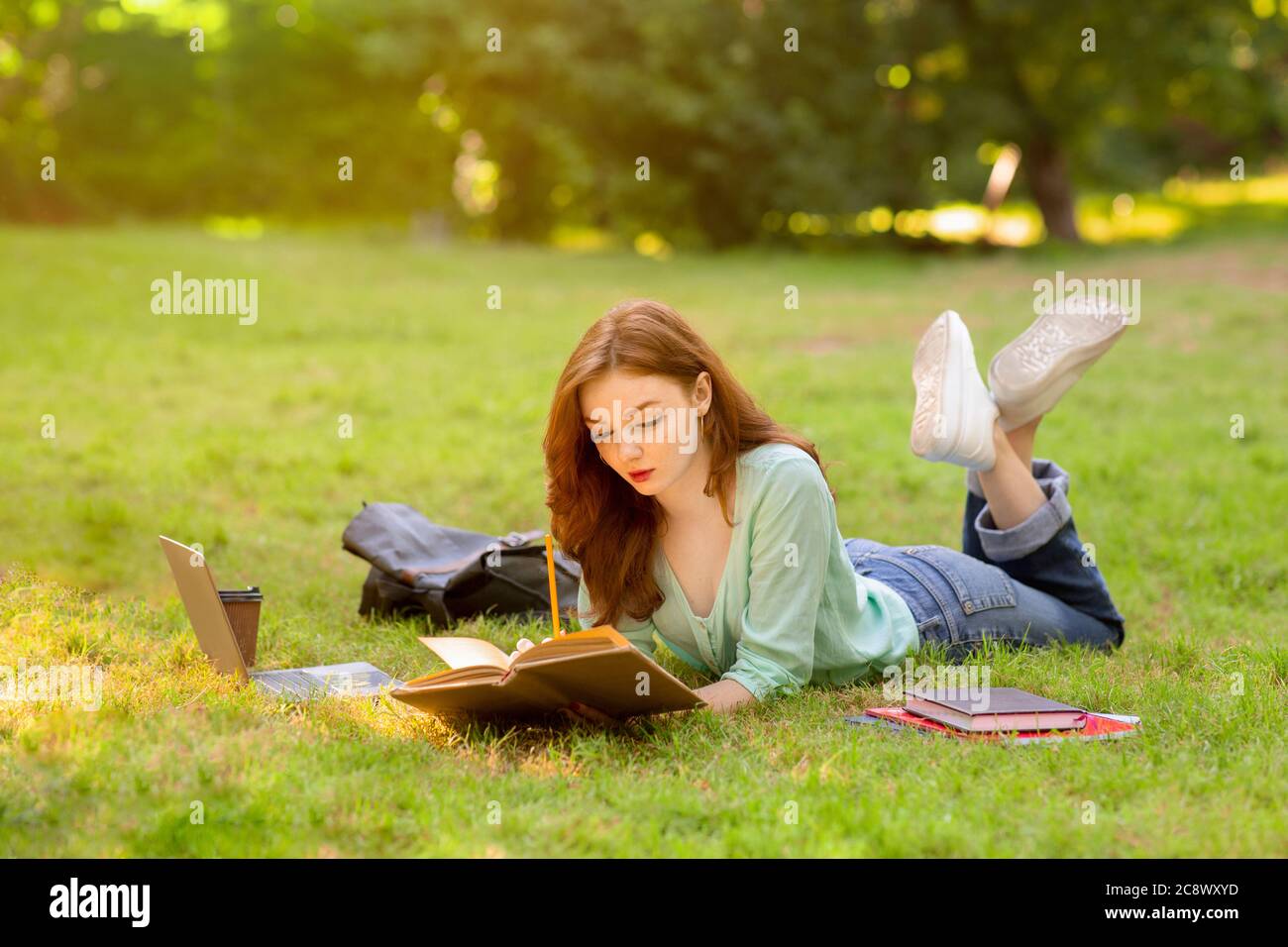 Beautiful student girl preparing for exams outdoors, lying on lawn with book Stock Photo