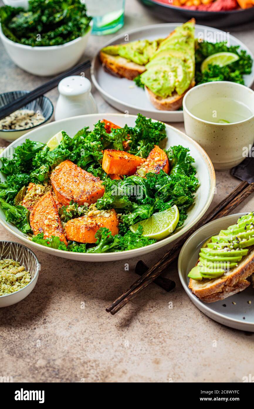 Vegan lunch table. Baked sweet potato salad with kale, avocado toast and hummus on a dark background. Stock Photo