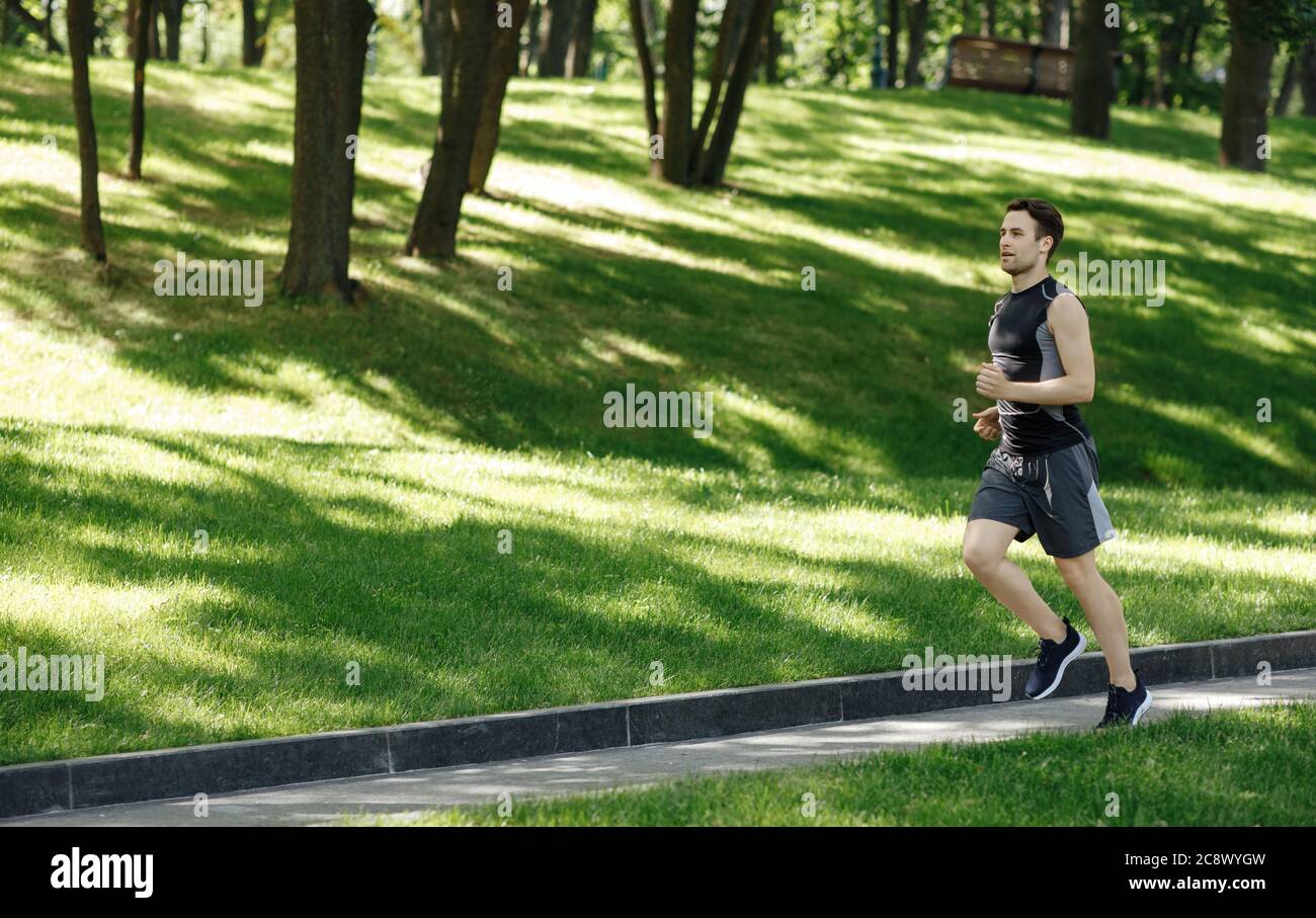 Physical activity outside. Guy with sneakers and fitness tracker runs at path in park Stock Photo
