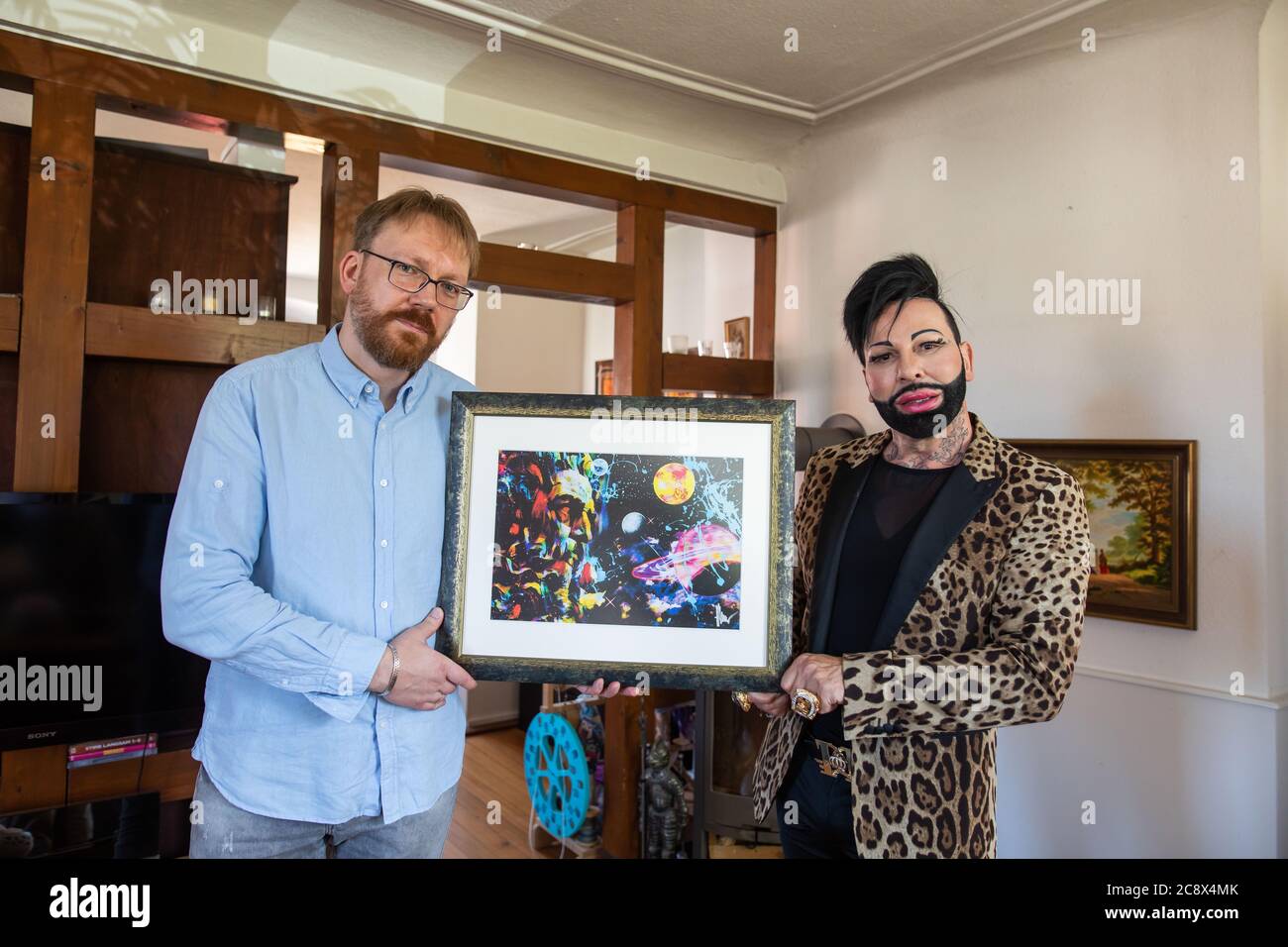 Rummingen, Germany. 27 July 2020, Baden-Wuerttemberg, Rümmingen: The protestant pastor Dirk Fiedler (l) and Harald Glööckler, fashion designer and entrepreneur, are standing in the protestant parsonage in Binzen and holding a picture of God that Glööckler has painted. Glööckler is to design a window which is to be given the place behind the altar in the church of Rümmingen, which has been walled up until now. Photo: Philipp von Ditfurth/dpa Credit: dpa picture alliance/Alamy Live News Stock Photo