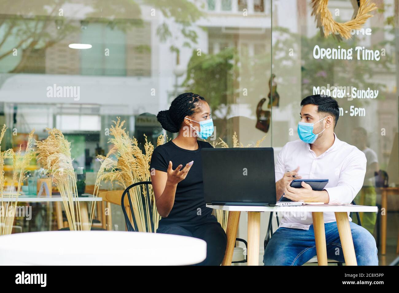 Multi-ethnic entrepreneurs in medical masks meeting at cafe table and discussing business strategy Stock Photo