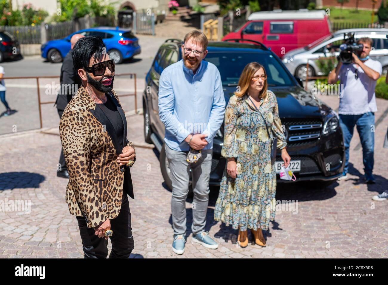 Rummingen, Germany. 27 July 2020, Baden-Wuerttemberg, Rümmingen: Harald Glööckler, fashion designer and entrepreneur (l), is standing in front of St. James' Church in Rümmingen with the pastors Dirk Fiedler and Christina Günther-Fiedler from the Lutheran parish of Binzen-Rümmingen. Glööckler is to design a window which will be given the place behind the altar in the church of Ruemmingen which has been walled up until now. Photo: Philipp von Ditfurth/dpa Credit: dpa picture alliance/Alamy Live News Stock Photo