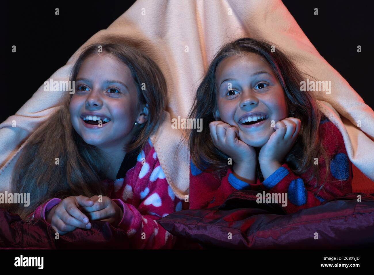 Children and fun time concept. Girls with excited faces. Girl friends watching TV in blanket tent. Kids wearing red jammies in bed on black background. PJs party for children. Stock Photo