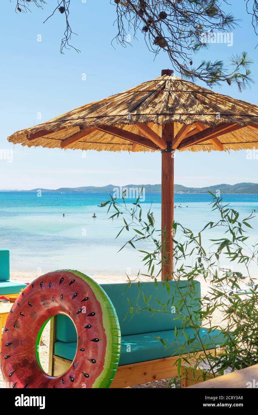 Wooden lounge with straw umbrella, bench and inflatable swim ring in the shade of pine tree on paradise beach looking out to the sea and blue sky. Stock Photo