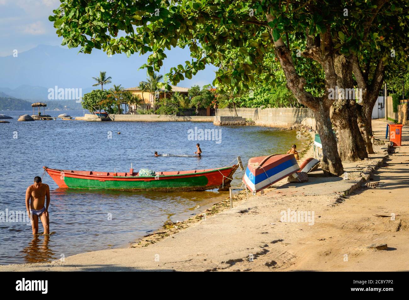 Locals bathing in the tranquil waters at Paquetá Island against lush trees, colorful fishing boats, and sunny sky. Stock Photo