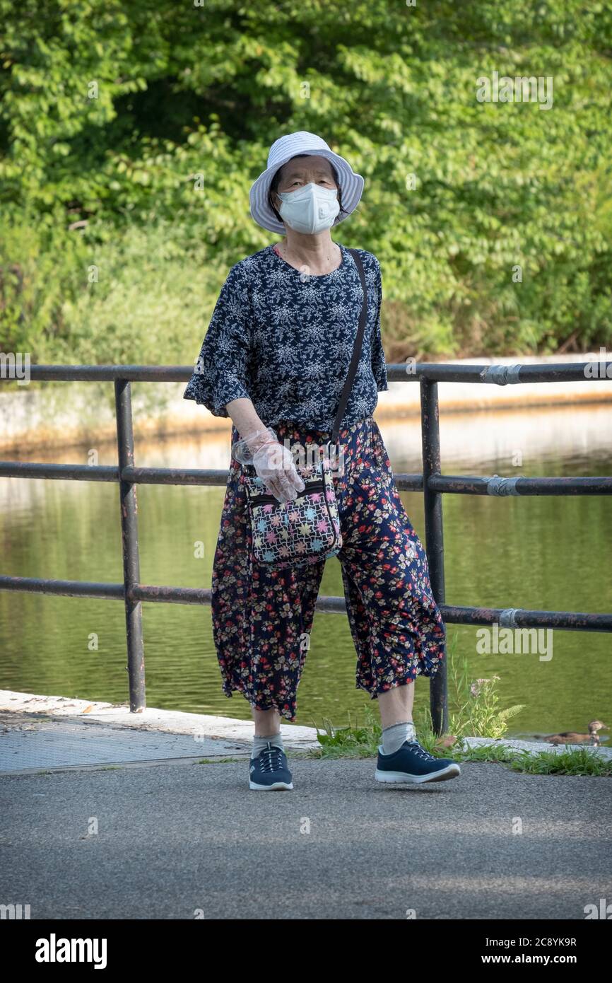 An older Asian American woman out for an exercise walk wearing a surgical mask & rubber gloves. In a park in Flushing, New York. Stock Photo