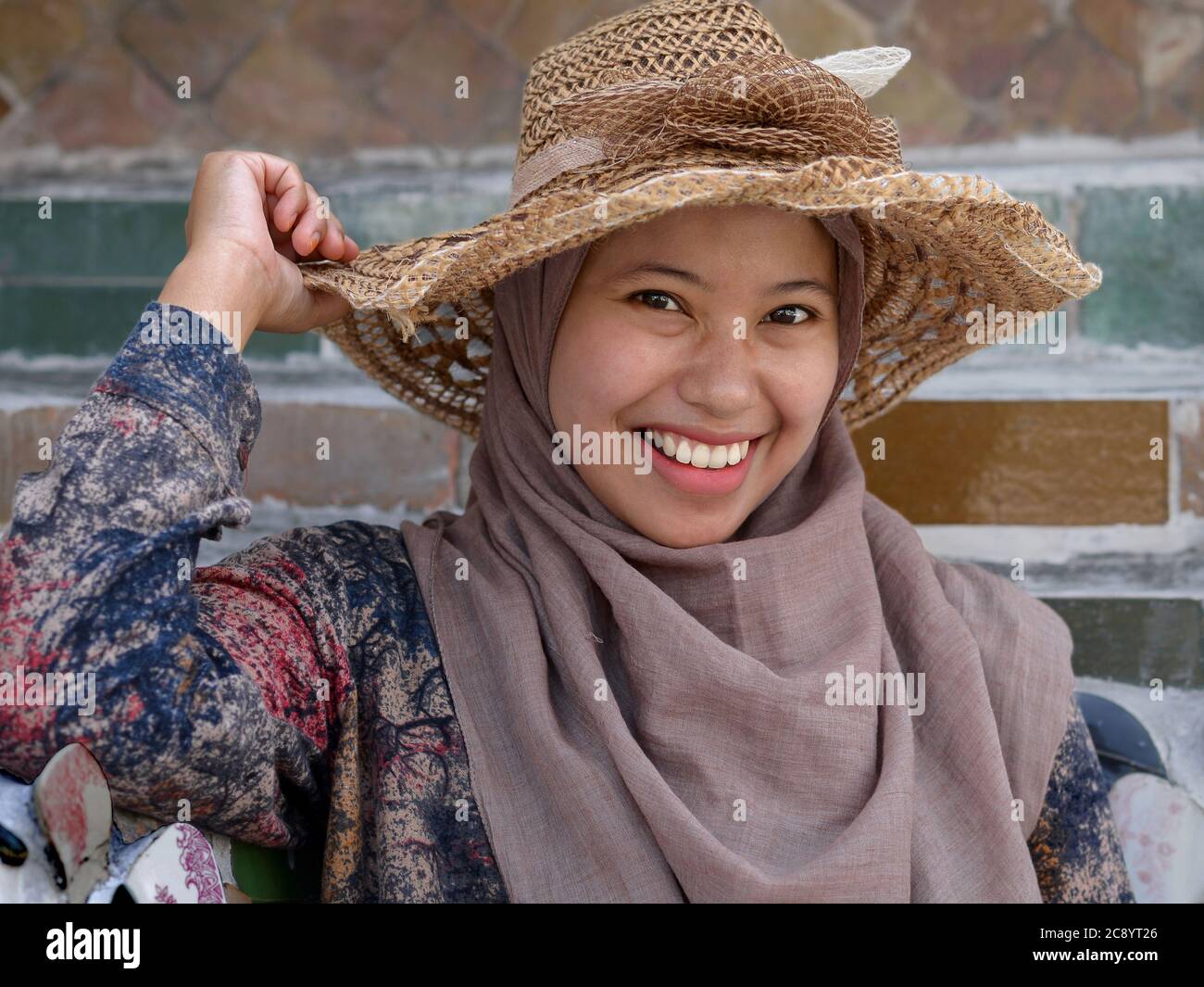 Young Indonesian woman wears a straw hat over her traditional hijab and smiles for the camera at Bangkok’s Wat Arun Temple. Stock Photo