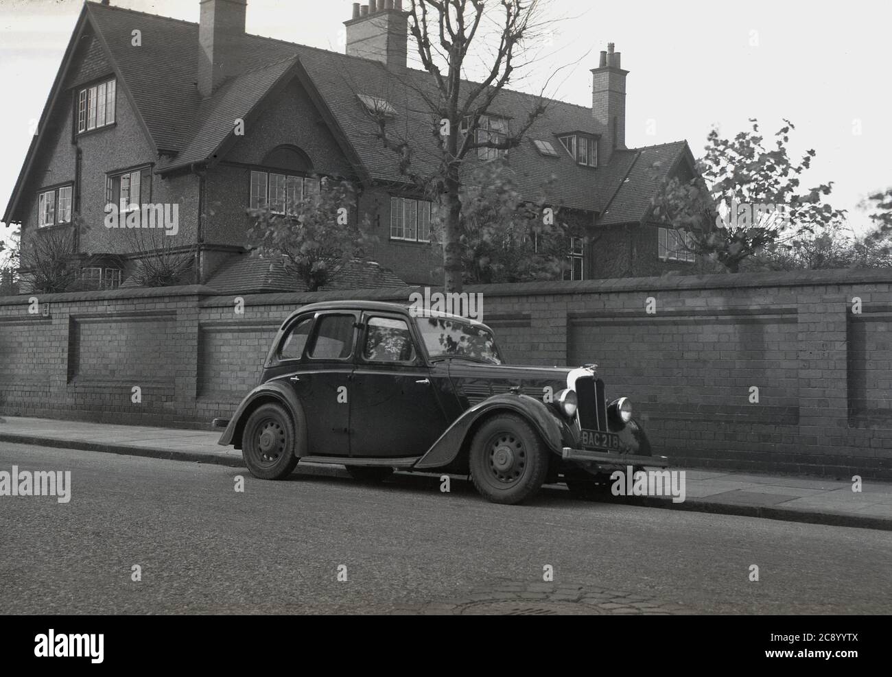 Circa 1940s, historical, a 4-door motorcar of the era, with its doors hinged in the middle, parked in a street beside a high brick wall surrounding a very large three-storey house with pebble-dash exterior, arts and craft in style, dating from the late 19th century, England, UK. The Arts and Crafts movement embraced architecture as a way to revive craftsmanship and individuality. Stock Photo
