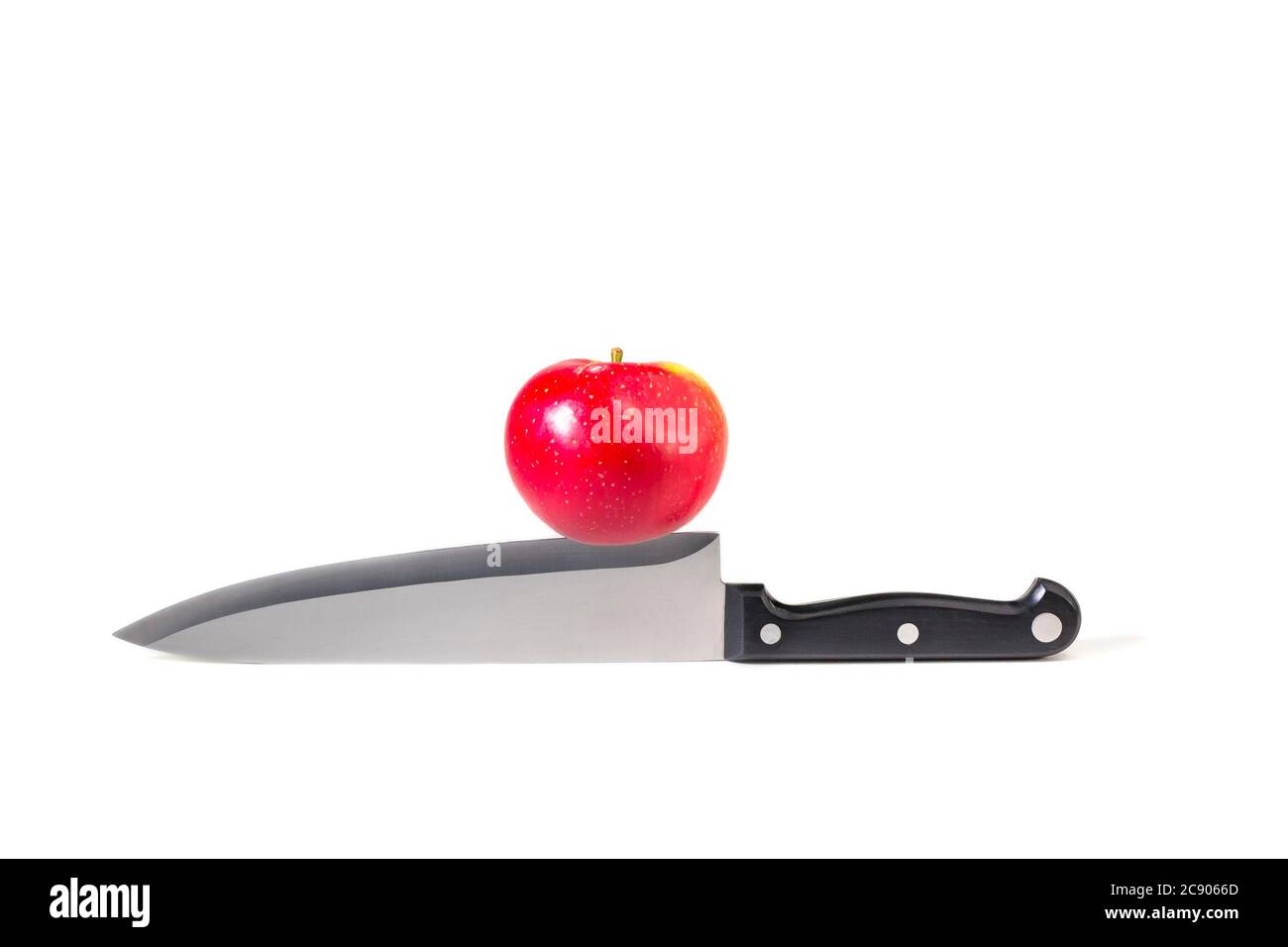 Red apple resting on cutting blade edge of large kitchen knife on white background. Concept photography: at the cutting edge, on a knife-edge. Stock Photo