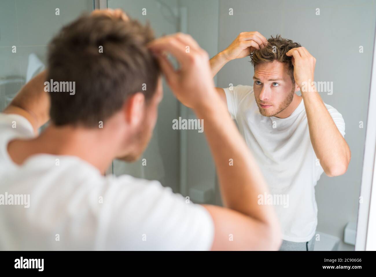 Hair loss man looking in bathroom mirror putting wax touching his hair styling or checking for hair loss problem. Male problem of losing hairs Stock Photo