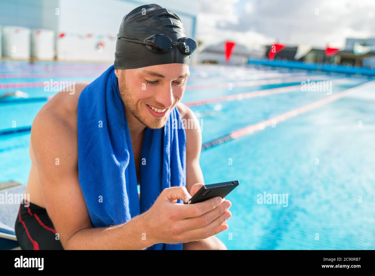 Swimmer man with swimming cap and goggles at pool using his mobile phone texting on smartphone app after training in outdoor pool. Happy athlete Stock Photo