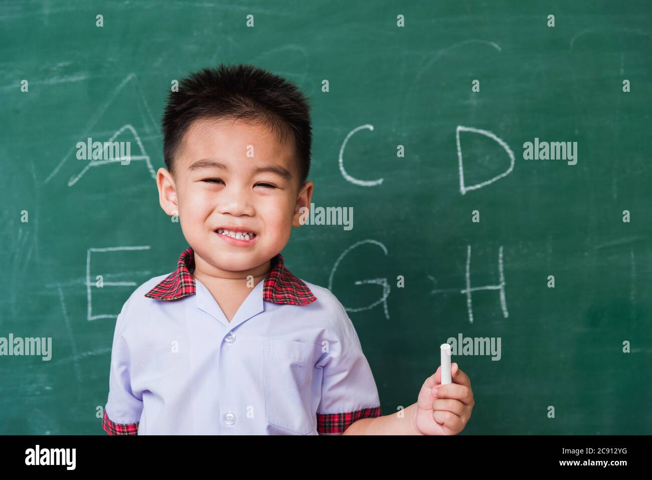 Back to School. Happy Asian funny cute little child boy kindergarten preschool smile in student uniform hold white chalk after write ABC with on green Stock Photo