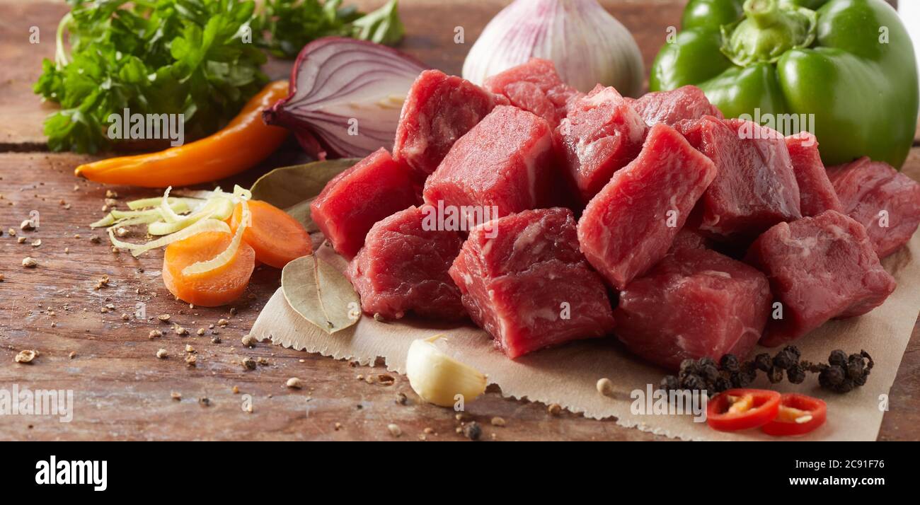 Diced fresh raw lean beef steak for goulash with an assortment of healthy vegetables, peppercorns, chili peppers and garlic in close up Stock Photo