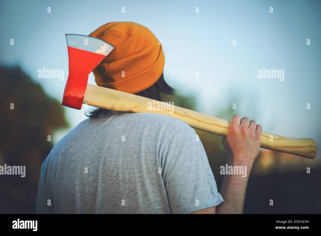 A young woodcutter in a yellow hat stands with a sharp red axe on his shoulder and looks dreamily at the sky. Stock Photo