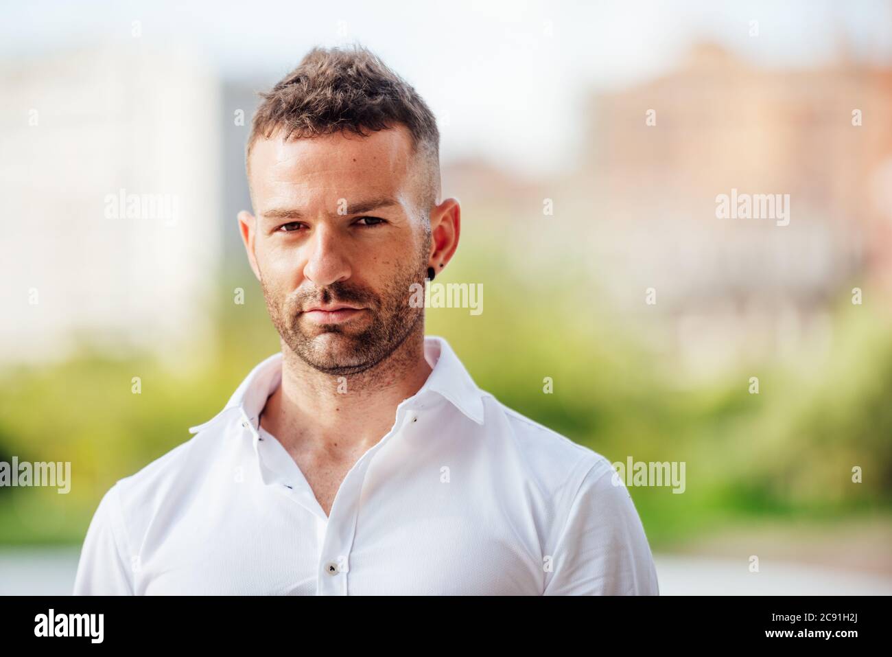 young man in white shirt looking at camera with short hair and short beard outdoors with plants and flowers background Stock Photo