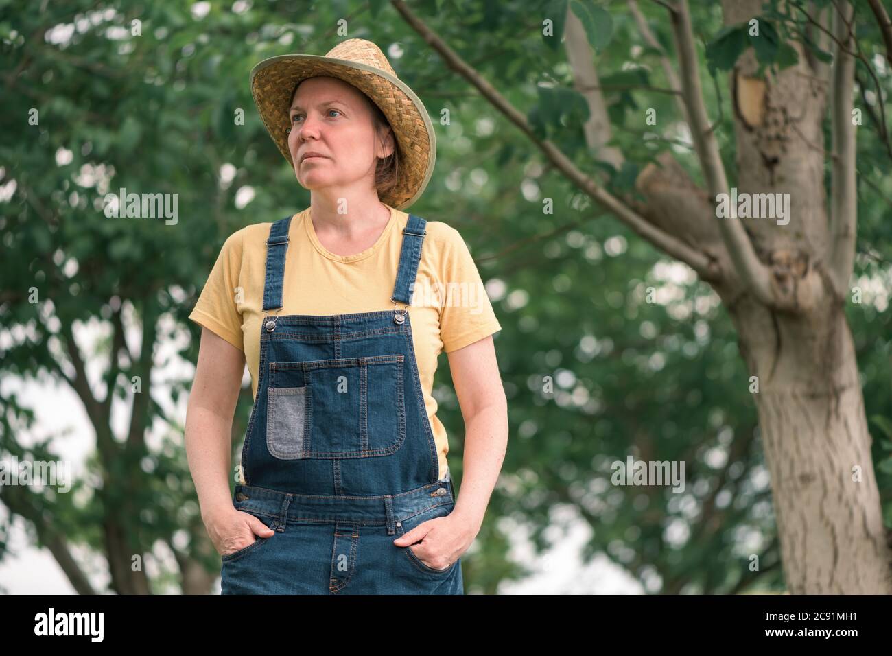 Portrait of female farmer posing in walnut fruit orchard, wearing straw hat and bib overalls Stock Photo