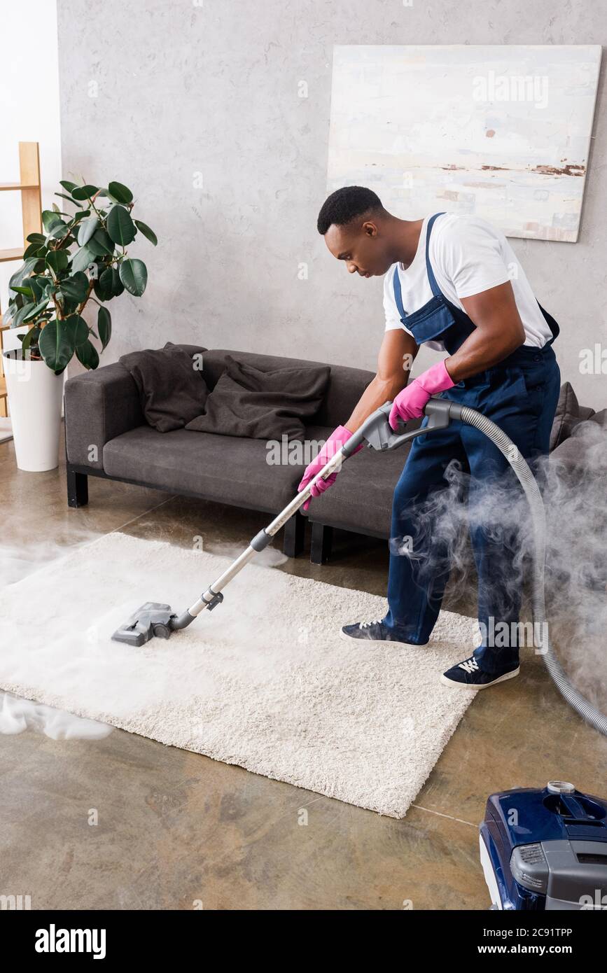 African american cleaner vacuuming carpet with hot steam in living room