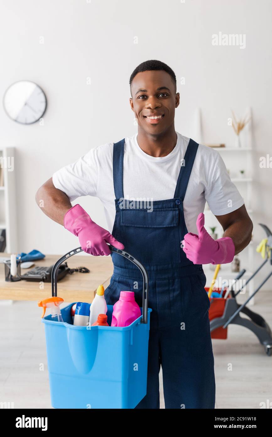 Smiling african american cleaner in uniform showing like and holding