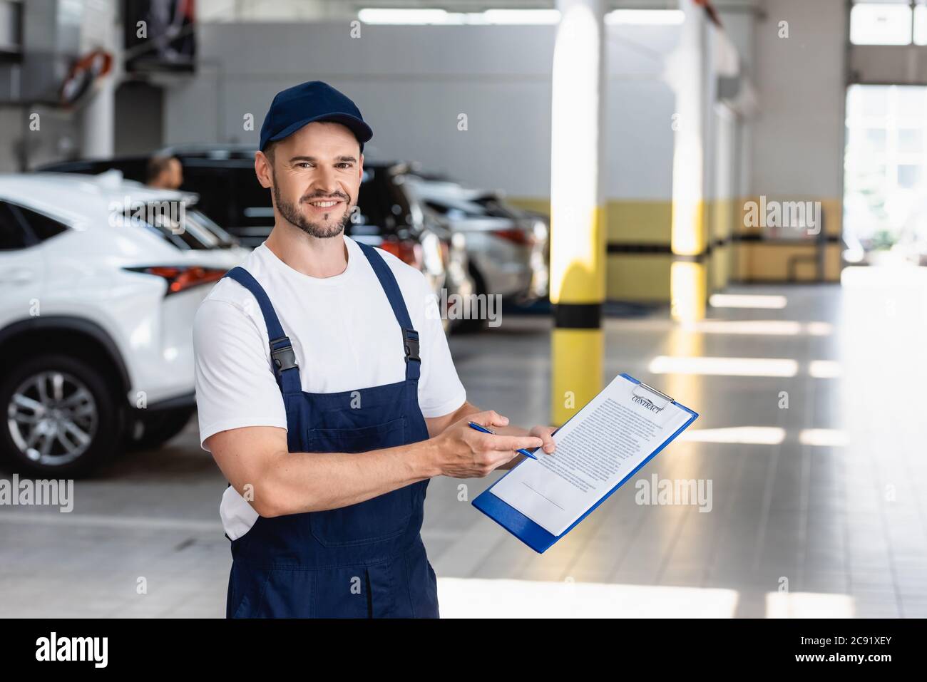 happy mechanic in uniform and cap holding clipboard with contract lettering and pen near cars Stock Photo