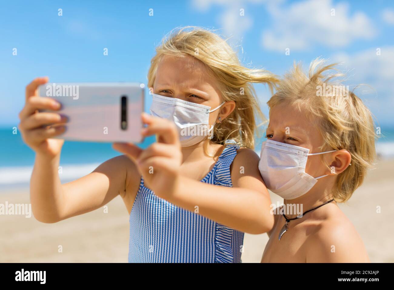 Funny kids taking selfie photo by smartphone on tropical sea beach. New rules to wear cloth face covering mask at public places due coronavirus COVID Stock Photo