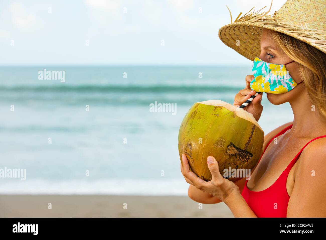 Funny portrait of woman in straw hat drinking young coconut on tropical sea beach.Rule to wear face  mask at public places due coronavirus COVID 19 Stock Photo