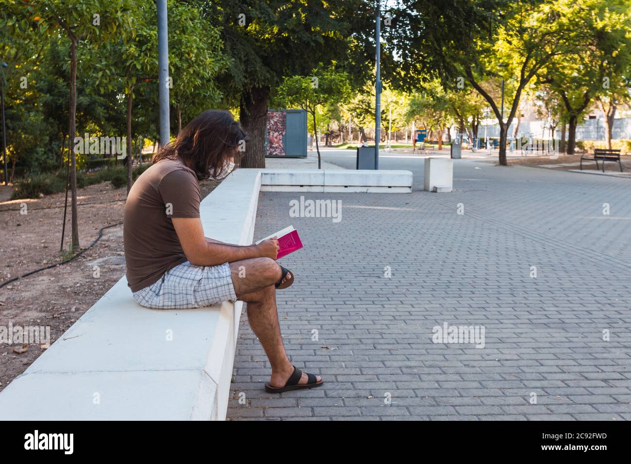 Male sitting on a stone bench in a park reading a book Stock Photo