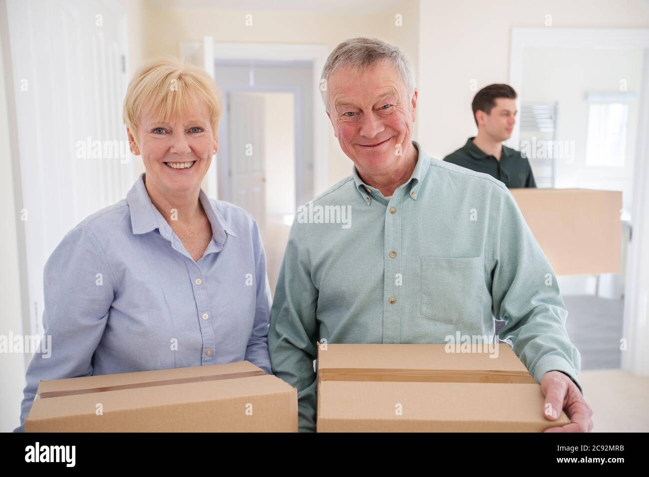 Portrait Of Senior Couple Downsizing In Retirement Carrying Boxes Into New Home On Moving Day With Removal Man Helping Stock Photo
