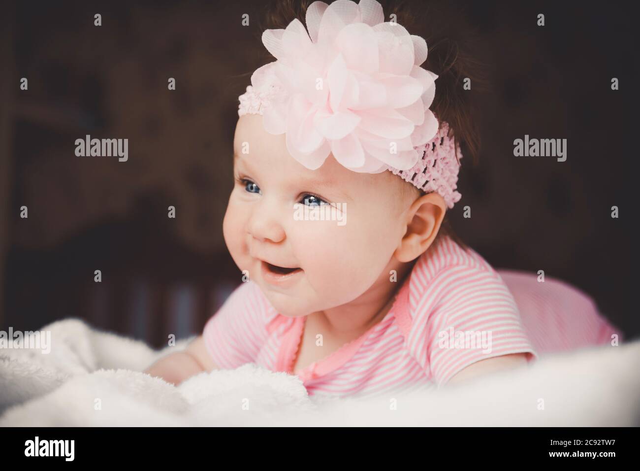 Close-up portrait of cute 3 months old smiling baby girl in pink lying down on a white bed at home. Big open eyes. Healthy little kid shortly after Stock Photo
