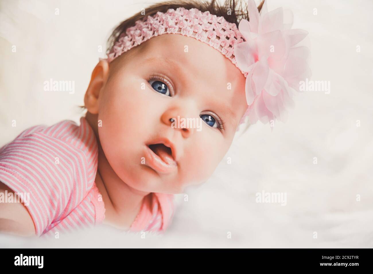 Close-up portrait of cute baby girl in pink lying down on a white bed. Looking at camera. Big open eyes. Healthy little kid shortly after birth. Stock Photo
