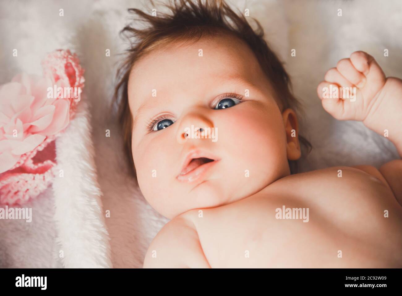 Close-up portrait of cute baby girl lying down on a white bed. Looking at camera. Big open eyes. Healthy little kid shortly after birth. Stock Photo