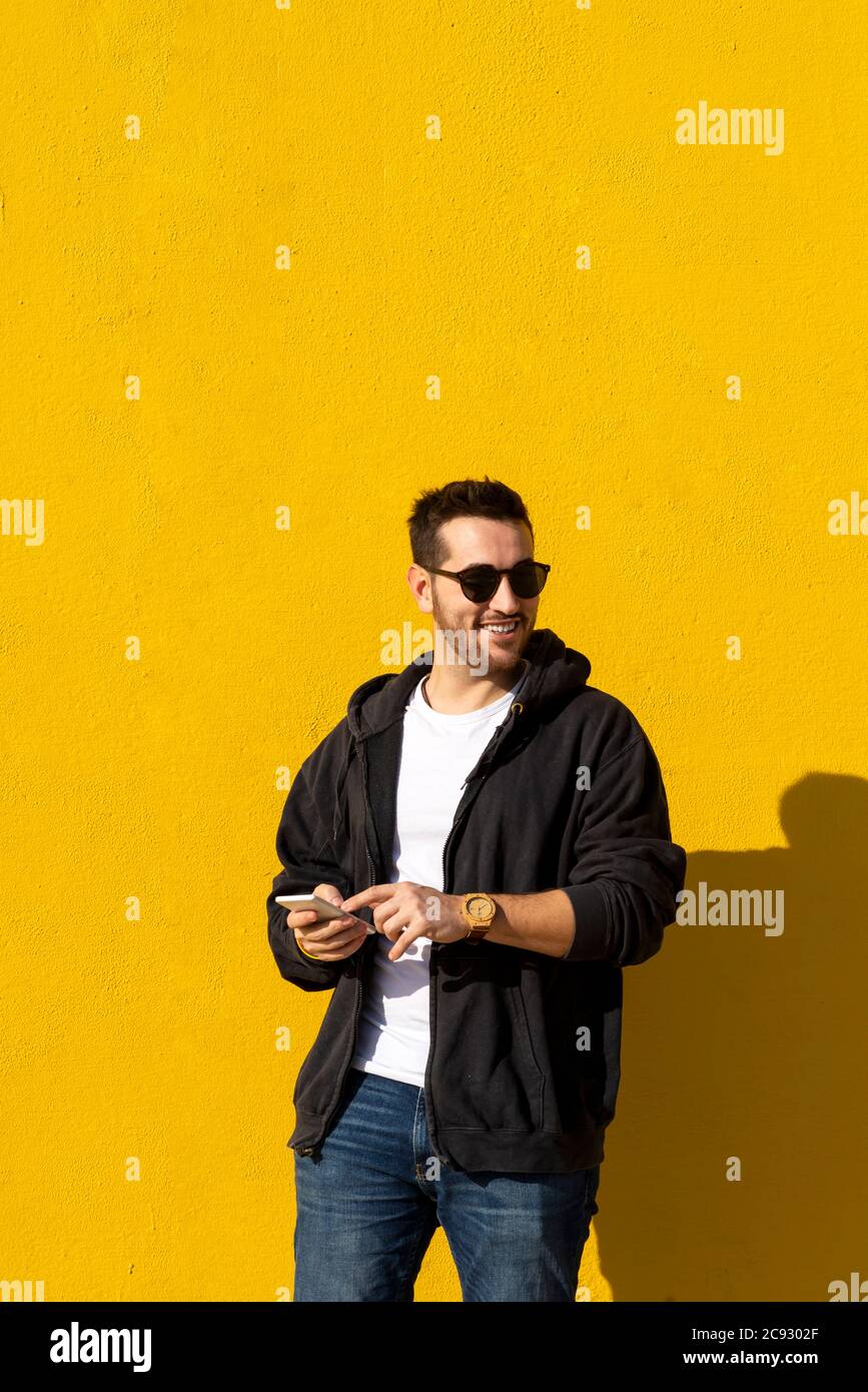 Bearded man with sunglasses standing against yellow wall, using phone Stock Photo