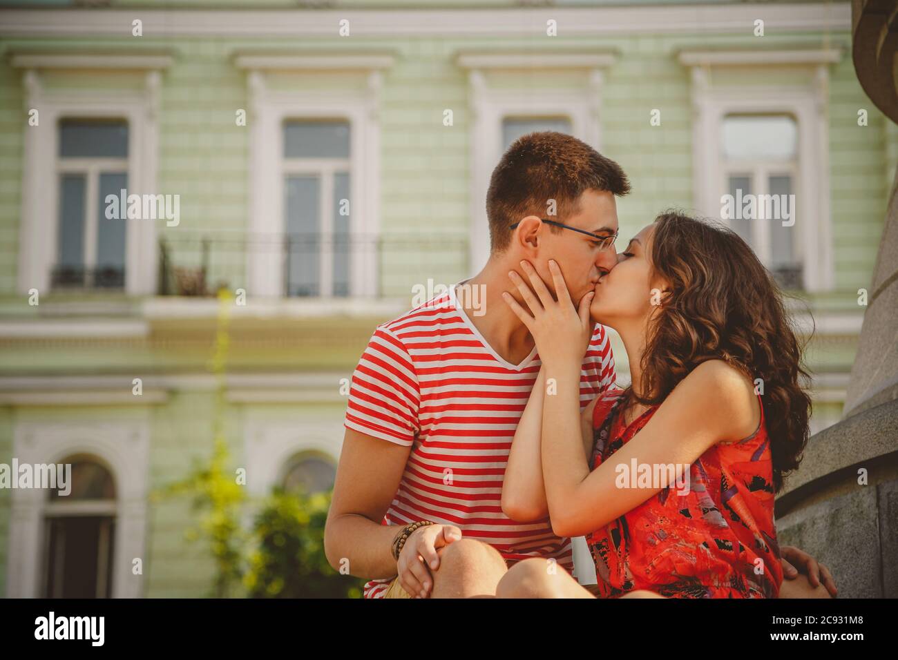 Beautiful young smiling couple in love kissing, hugging, sitting outdoors at green city street, summertime Stock Photo