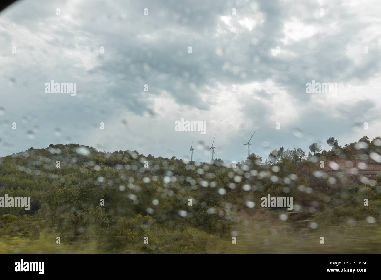 Wind turbines under a cloudy sky seen through the wet glass window of ...