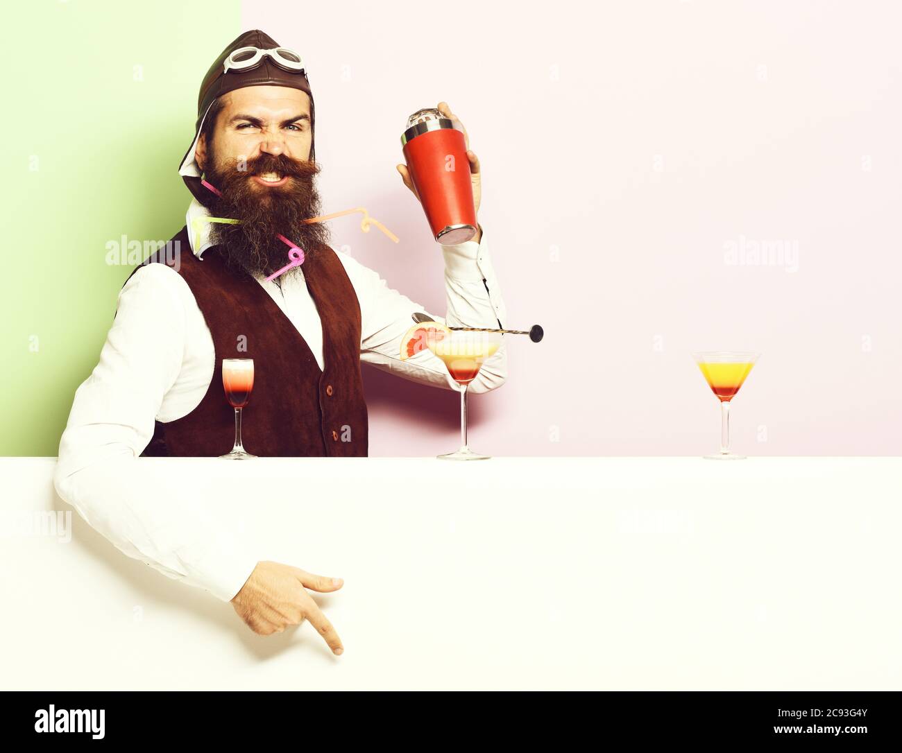 handsome bearded pilot with long beard and mustache on funny face with shaker alcoholic cocktails in vintage suede leather waistcoat with hand and glasses, on purple green studio background Stock Photo
