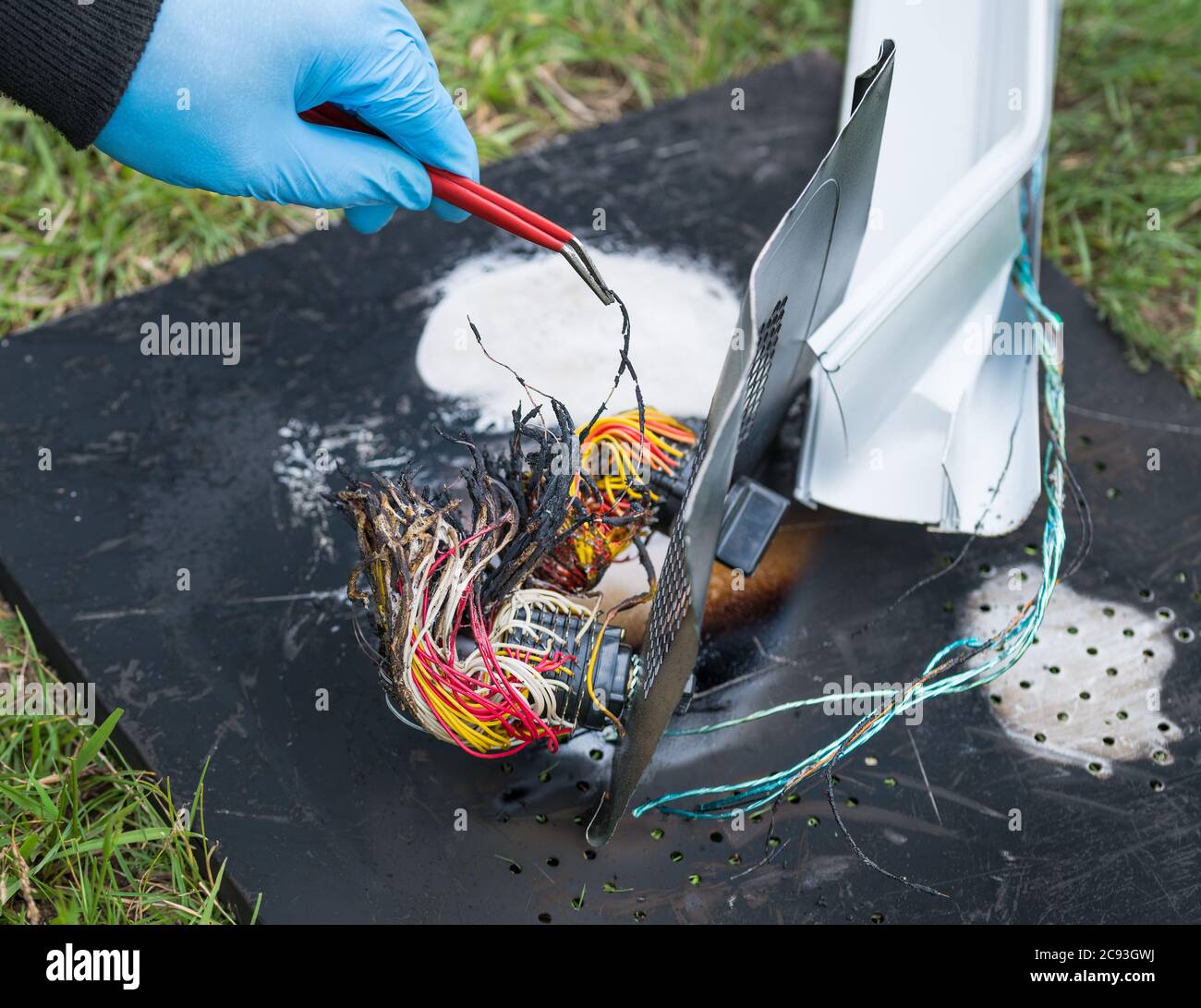 Hand in protective glove holding damaged electric wires with tweezers. Detective work on charred wreck in grass. Road, rail or air crash investigation. Stock Photo