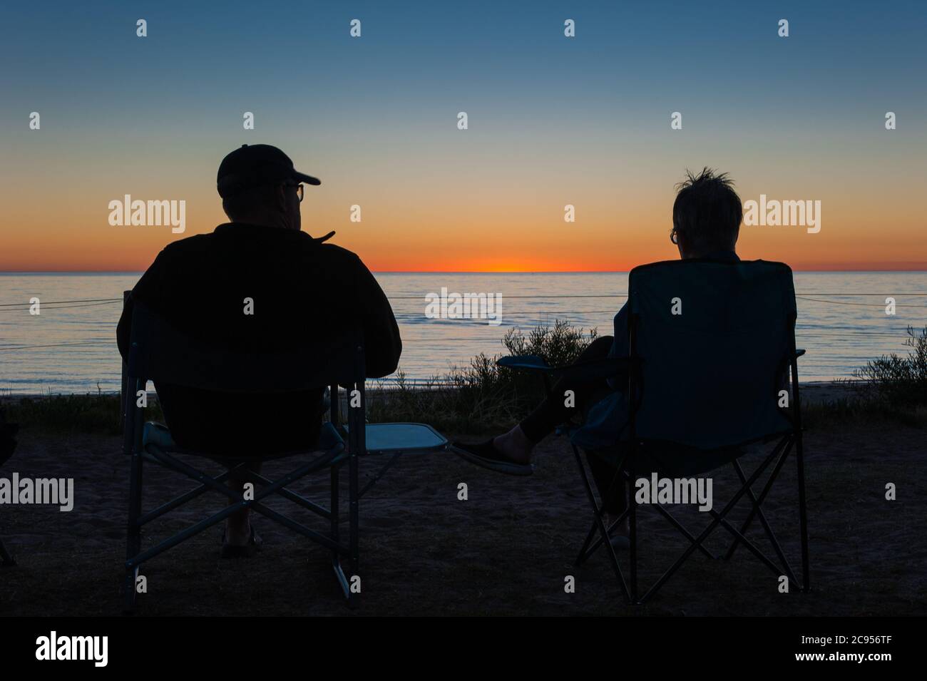 A tourist couple in silhouette sit enjoying happy hour and a vibrant sunset facing the southern ocean at Brighton Beach, South Australia. Stock Photo