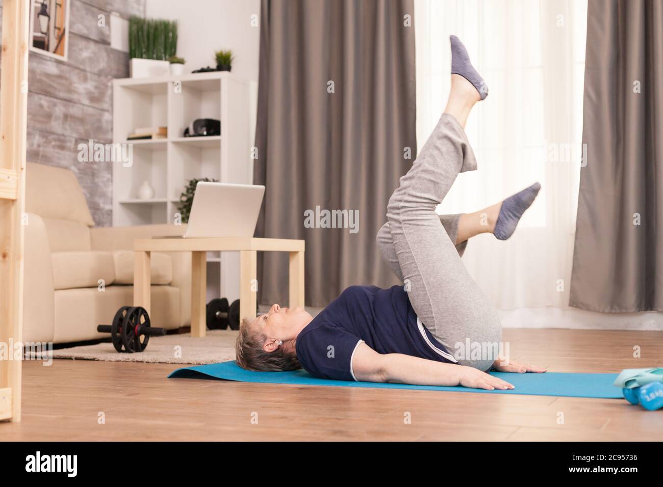 Old woman exercising in her apartment sitting on yoga mat. Old person  pensioner online internet exercise