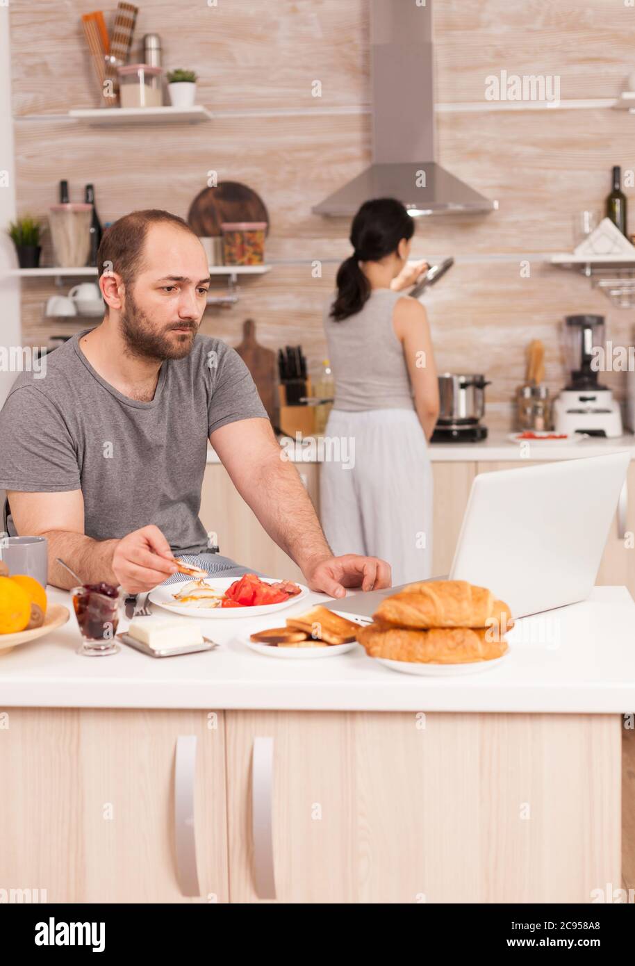 Entrepreneur working from home while eating breakfast in kitchen, wearing pajamas enjoying roasted bread with butter. Freelancer working online via internet using modern digital technology Stock Photo