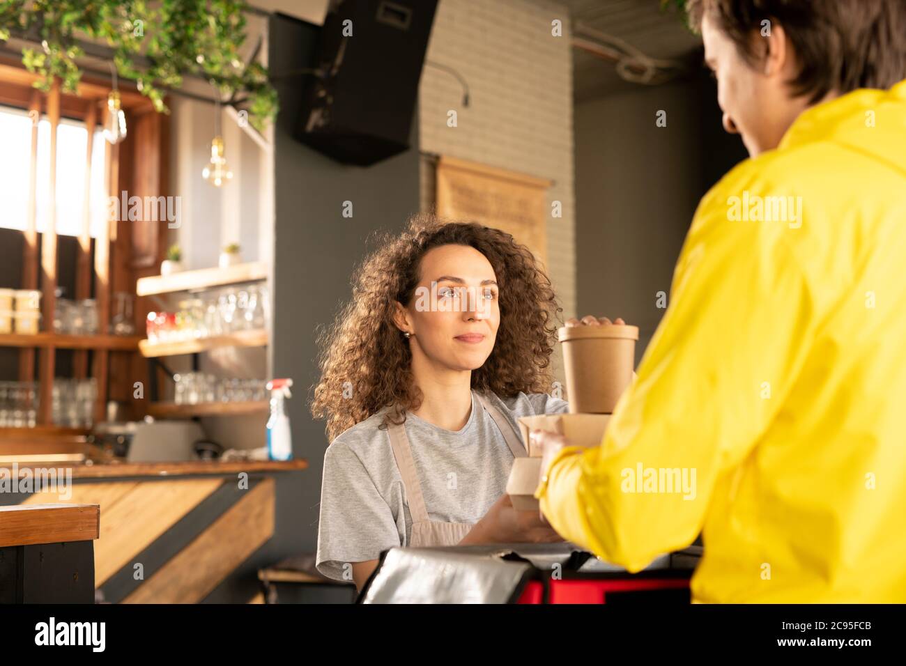 Content pretty waitress with curly hair giving packaged boxes to deliver boy for delivering to customer during coronavirus epidemic Stock Photo