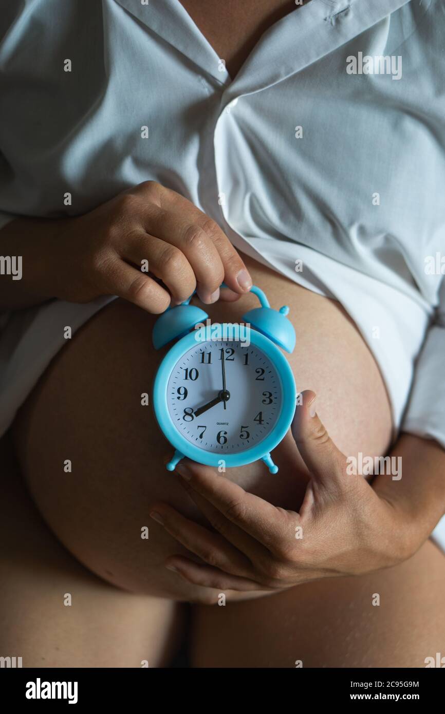 High angle of crop pregnant female holding alarm clock on belly while waiting for baby. Stock Photo