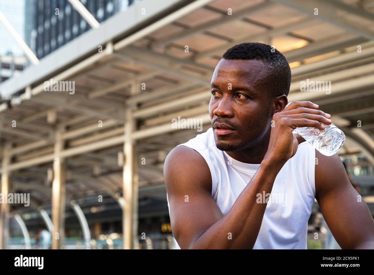 portrait of relaxing young african american sportsman sitting and drinking water from bottle after running exercise in city in morning with building b Stock Photo