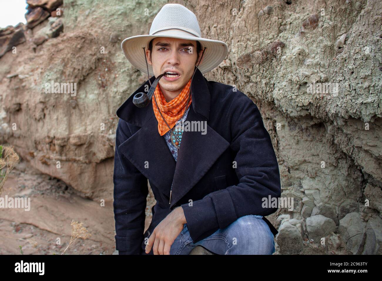 Serious cowboy wearing a wide brimmed hat and bandanna smokes a tobacco pipe in an arid desert canyon in the Ojito Wilderness, New Mexico Stock Photo