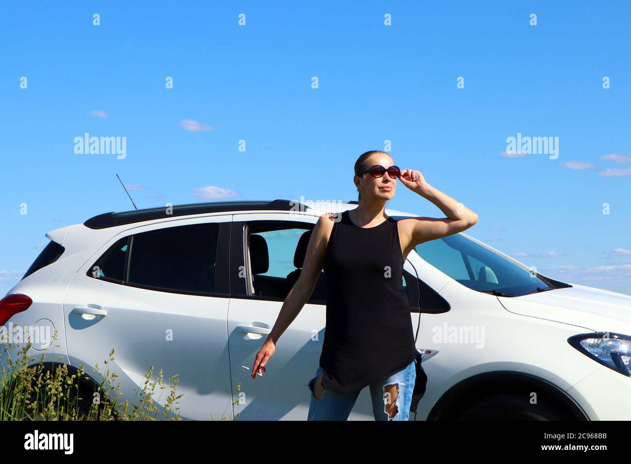 Pretty young woman wearing sunglasses standing near the white car against bright blue sky Stock Photo
