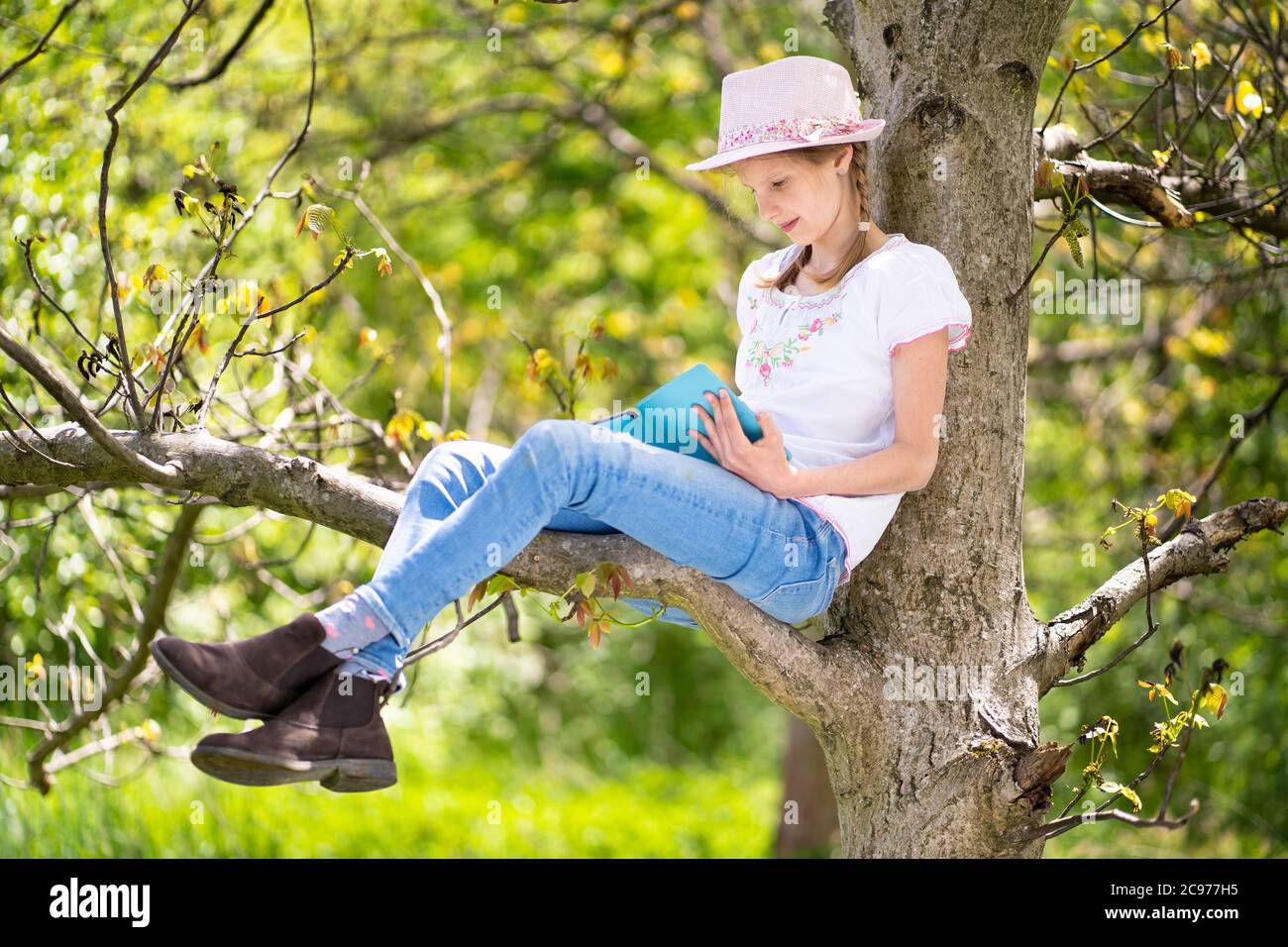 Teenage girl in white T-shirt and pink hat is sitting on a tree in the garden, smiling and reading book on ebook reader. Great idea to spend free time. Stock Photo