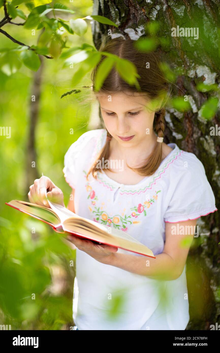 Teenage girl in white T-shirt standing by the tree in the forest and reading interesting book. Great idea and activity to spend summer holidays. Stock Photo