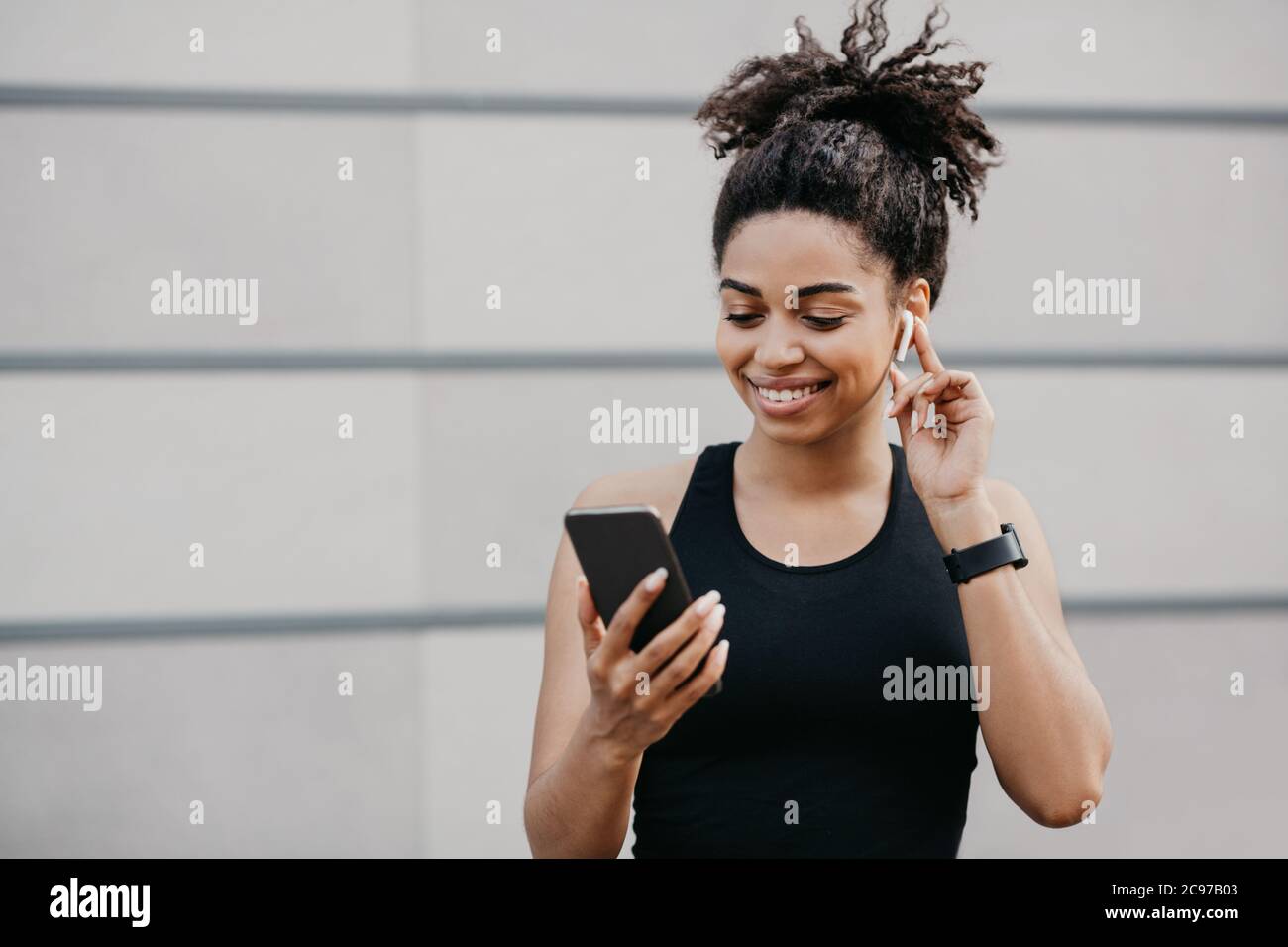 Smiling african american woman with fitness tracker, puts on wireless headphone and choosing song in smartphone Stock Photo