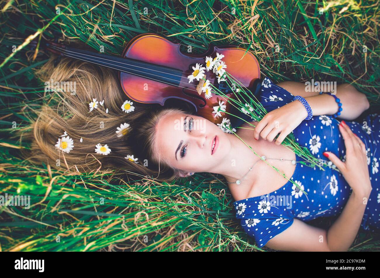 Young slim fair-skinned girl blonde beautiful lies in tall grass. Top view. Girl in a blue short tight dress with a print of daisies. A bouquet and lo Stock Photo