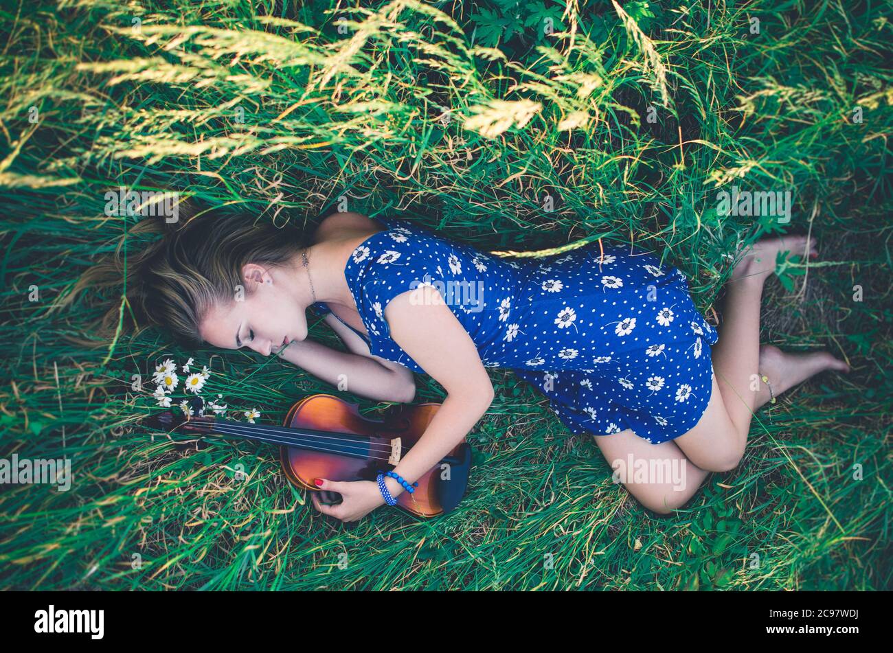 Young slim fair-skinned girl blonde lies in tall grass. Top view. Girl in a blue short tight dress with a print of daisies. Violin Stock Photo