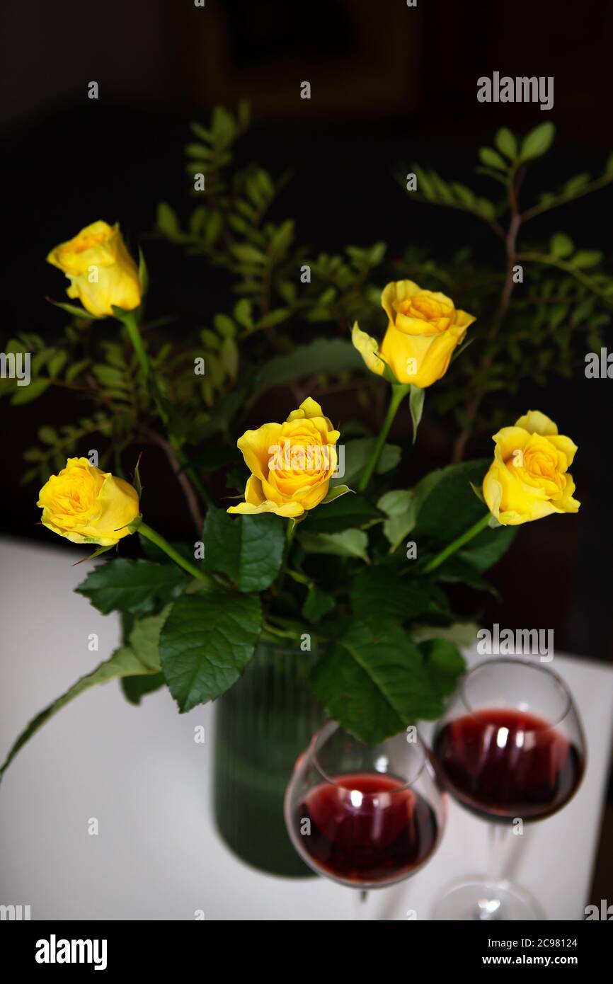 A very beautiful bouquet of yellow roses stands in a green vase and two glasses of red wine. Romantic evening Stock Photo