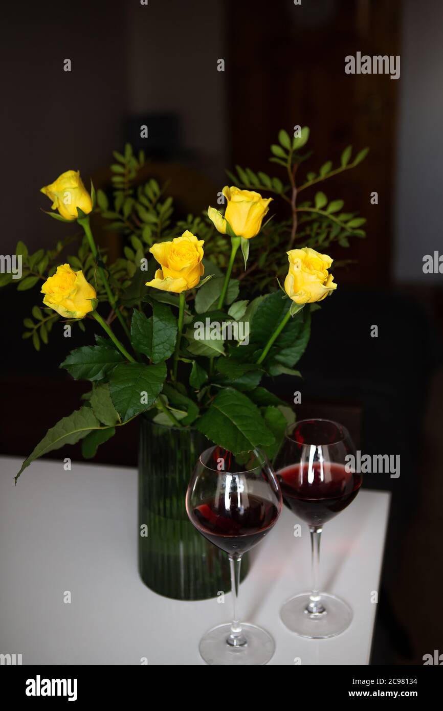 A very beautiful bouquet of yellow roses stands in a green vase and two glasses of red wine. Romantic evening Stock Photo